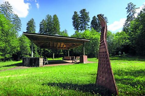 Skulptur aus Ton in Form einer stilisierten Welle, im Vordergrund auf einer grünen Wiese, im Hintergrund ein Pavillon mit Sitzgelegenheiten und Bäumen.