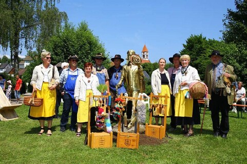 Gruppe von Menschen in traditioneller Kleidung, die auf einer Wiese steht. Einige halten Körbe, während andere eine goldene Statue und dekorative Elemente präsentieren. Heller Himmel und grüne Bäume im Hintergrund.