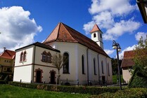 Ansicht einer weißen Kirche mit rotem Satteldach und dazugehörigem Gebäude. Umgeben von grünem Rasen und Bäumen unter blauem Himmel mit Wolken.