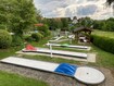 Minigolfplatz mit mehreren Bahnen, umgeben von Bäumen und Sträuchern, im Hintergrund ein Pavillon.